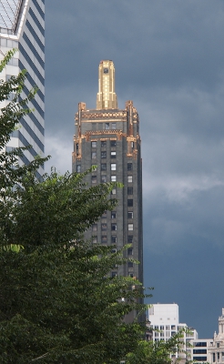 [Against the dark blue-grey skies of an impending storm is a grey building with many gold highlights around the edges of the top and with a gold tower on the roof.]